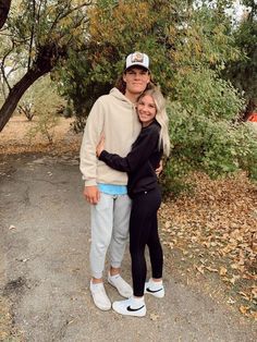 a man and woman standing next to each other in front of some trees with leaves on the ground