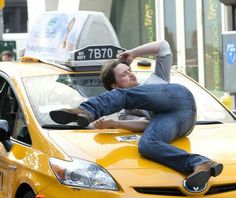 a man laying on top of a yellow car in the middle of a busy street