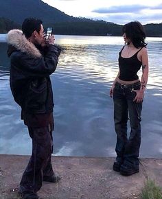 a man and woman standing next to each other by the water taking pictures with their cell phones