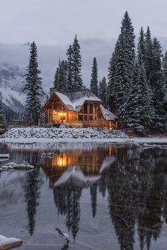 a cabin sits on the shore of a lake in front of snow covered evergreen trees