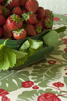 strawberries in a green bowl on a tablecloth with red flowers and leaves next to it