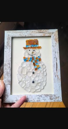 a hand holding up a white frame with a snowman made out of pebbles in it