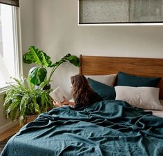 a woman laying in bed reading a book next to a potted plant and window