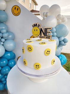 a white cake sitting on top of a table covered in blue and yellow balloons with smiley faces