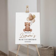 a white sign with a teddy bear holding balloons on it's easel for a first birthday party