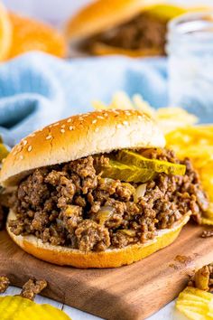 a sloppy joe sandwich on a cutting board with pickles and mustard in the background