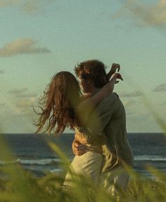 a man holding a woman in his arms while standing on the grass by the ocean