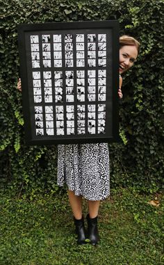 a woman standing in front of a hedge holding up a black and white framed poster