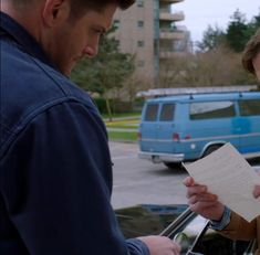 two men standing next to each other in front of a blue van and one holding a piece of paper