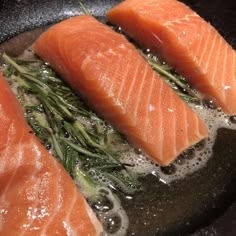 three pieces of salmon are being cooked in a skillet with water and herbs on the side