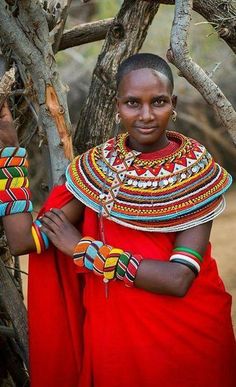 a woman in a red dress with bracelets and necklaces on her neck standing next to a tree