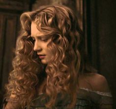 a woman with long curly hair standing in front of a door and looking off to the side