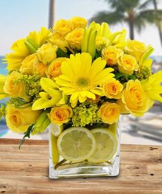 a vase filled with yellow flowers and lemons on top of a wooden table next to the ocean