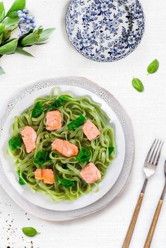 a white plate topped with pasta covered in salmon and greens next to utensils