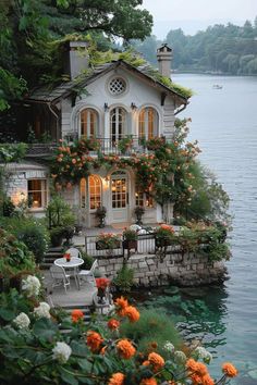 the house is surrounded by flowers and greenery on the water's edge, with boats in the background