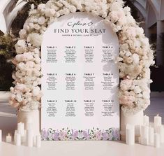 a wedding seating chart with white flowers and candles on the table in front of an arch