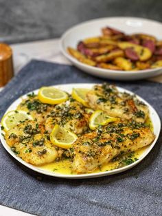 a white plate topped with chicken covered in lemons and parsley next to a bowl of potatoes