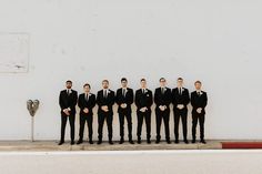 a group of men standing next to each other in front of a white wall wearing black suits