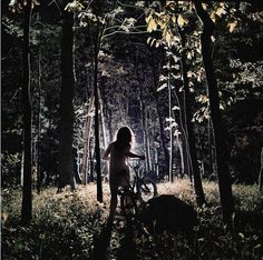 a woman standing in the woods next to her bike with trees around her and light coming from behind