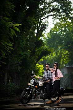 two people are sitting on a motorcycle in the middle of the road with trees behind them