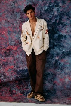 a man standing in front of a wall wearing a white blazer and brown pants