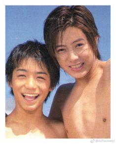 two young men standing next to each other near the ocean smiling at the camera, with one man's shirtless torso visible