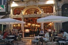 people sitting at tables in front of a building with white umbrellas on the outside