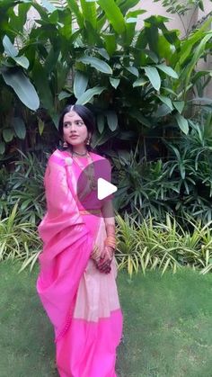 a woman in a pink and beige sari standing on grass with plants behind her