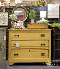 a yellow dresser with some plants on top and a mirror above it in a store