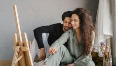 a man and woman sitting on the floor next to an easel