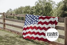 an american flag quilt hanging on a fence in front of a field with trees and grass