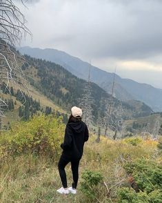 a woman standing on top of a grass covered hillside next to trees and mountains in the distance