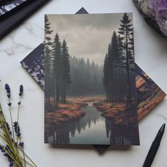 three cards with trees and water on them sitting next to some rocks, pens and flowers