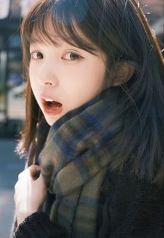 a young woman with long hair wearing a scarf and looking at the camera while standing in front of a building