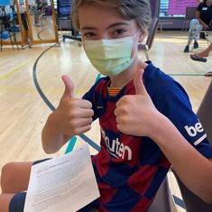 a young boy wearing a face mask and giving the thumbs up while sitting on a basketball court