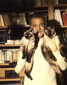 a man holding two siamese cats in front of his face and looking at the camera