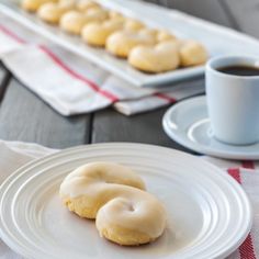 two glazed donuts on a white plate next to a cup of coffee and tray of cookies