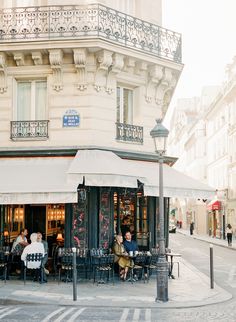 people are sitting at tables in front of a building on a street corner with white awnings