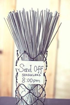 a glass vase filled with lots of sticks on top of a wooden table next to a sign that reads send off sparklers 3 00pmm