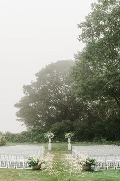 an outdoor ceremony set up with white chairs and flower arrangements on the grass in front of trees