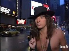 a man with long hair wearing a fedora in the middle of a city at night