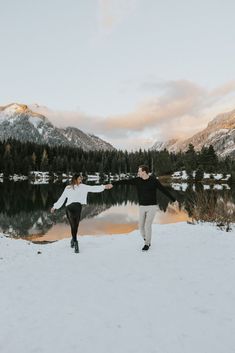 a man and woman holding hands in the snow by a lake with mountains in the background