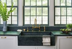 a kitchen sink with green counter tops and white cabinets in front of two large windows