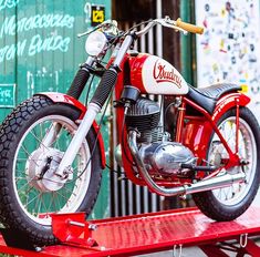 a red motorcycle sitting on top of a wooden table next to a green building with writing on it