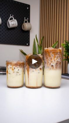 there are three jars with food in them on the counter next to a plant and coffee mugs