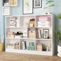 a white book shelf filled with books next to a potted plant and pictures on the wall