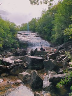 people are sitting on rocks in the water near a small waterfall and some green trees