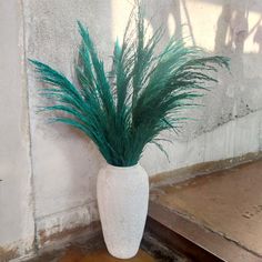 a white vase filled with green grass on top of a wooden floor next to a wall