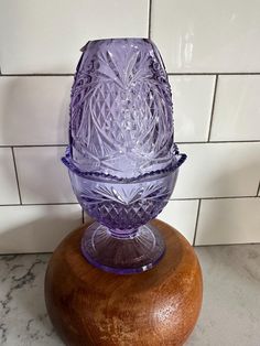 a purple glass vase sitting on top of a wooden stand in front of a white tile wall