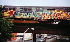 a train covered in graffiti on top of a bridge over a street with cars parked below